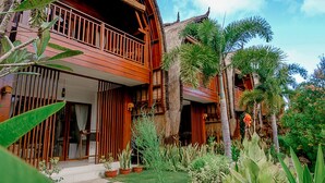 Una casa de madera de dos pisos con un balcón, rodeada de plantas tropicales y un camino de piedra.