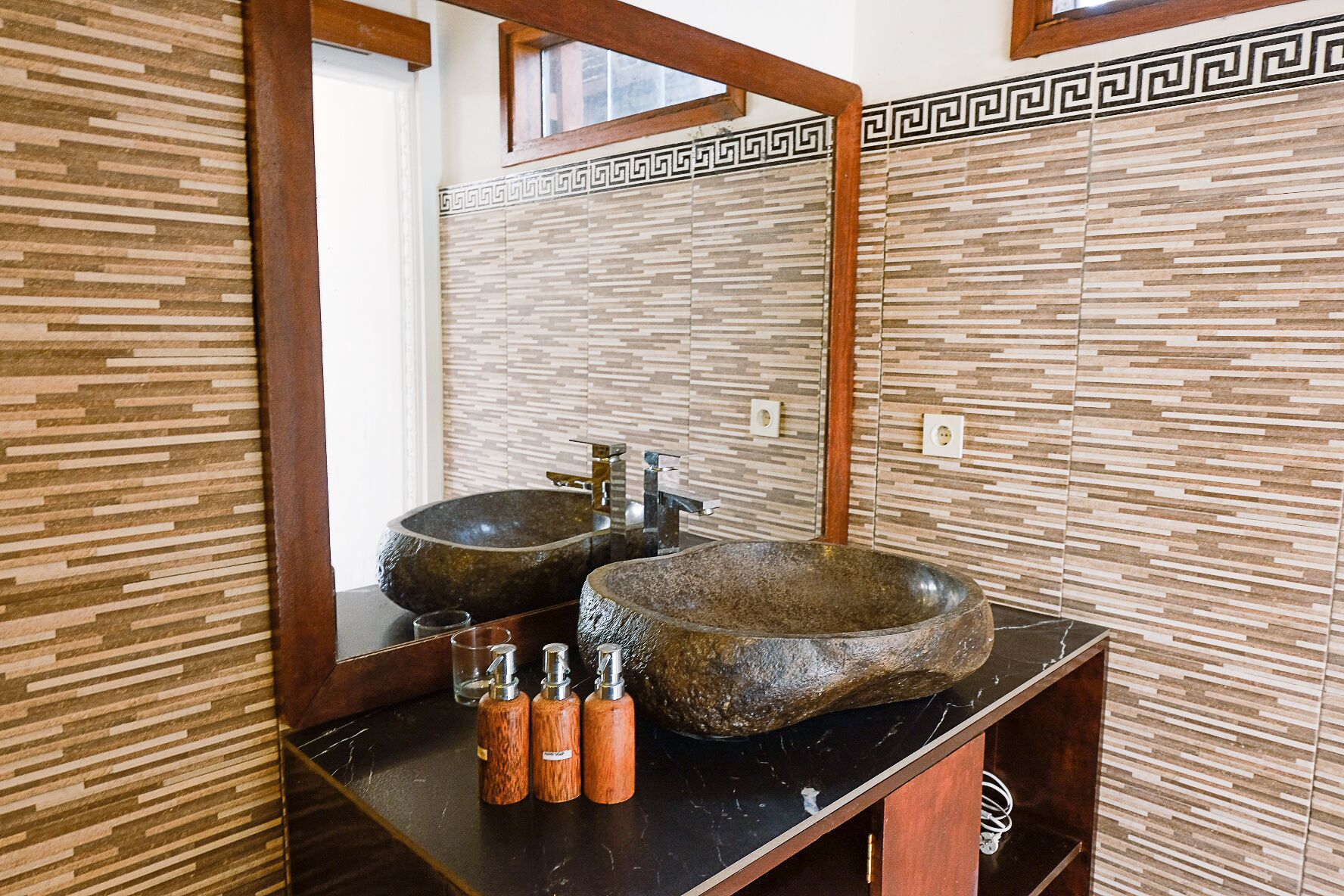 A bathroom with two stone sinks, a wooden vanity, and a mirror with a geometric border.