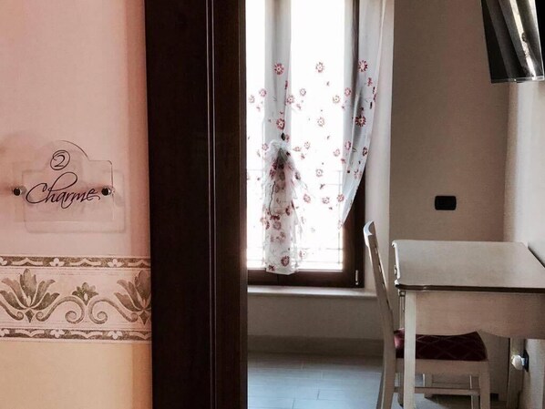 A room with a dining table and chairs, a window with floral curtains, and a wall with a decorative border and a sign that reads "Charmes".