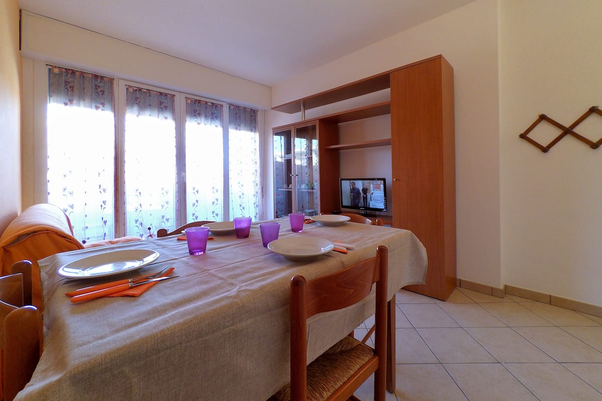A dining area with a table set for four, a television on a stand, and a wooden shelf unit against the wall.