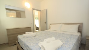Ein ordentlich bezogenes Bett mit einer blauen Bettdecke und zwei weißen Kissen. Ein Holznachttisch mit einer Lampe steht daneben, und über dem Bett befindet sich ein Spiegel an der Wand.