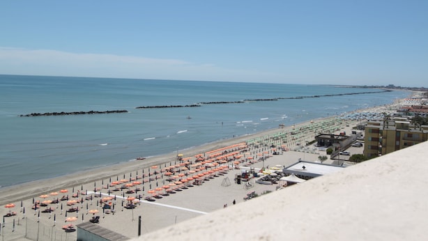 Ein Strand mit mehreren Reihen von Sonnenschirmen und Liegestühlen, einer Promenade und Gebäuden im Hintergrund.