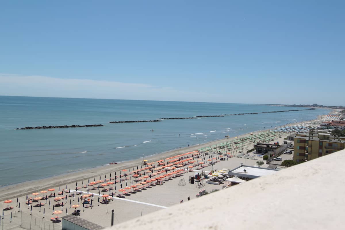 A beach with rows of umbrellas and sun loungers, a promenade, and buildings in the background.