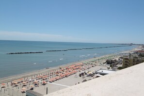 A beach with rows of umbrellas and sun loungers, a promenade, and buildings in the background. - Eco del Mare Apartments (Comacchio)