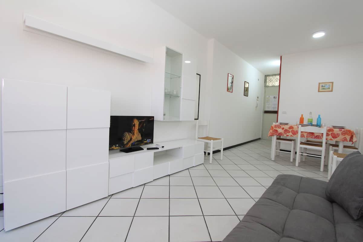 A modern living room with a flat-screen TV, white cabinetry, a gray sofa, and a dining area with a table and chairs.