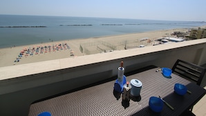 Ein Balkon mit Esstisch, der auf einen Strand mit Sonnenschirmen und freie Meerblick blickt.