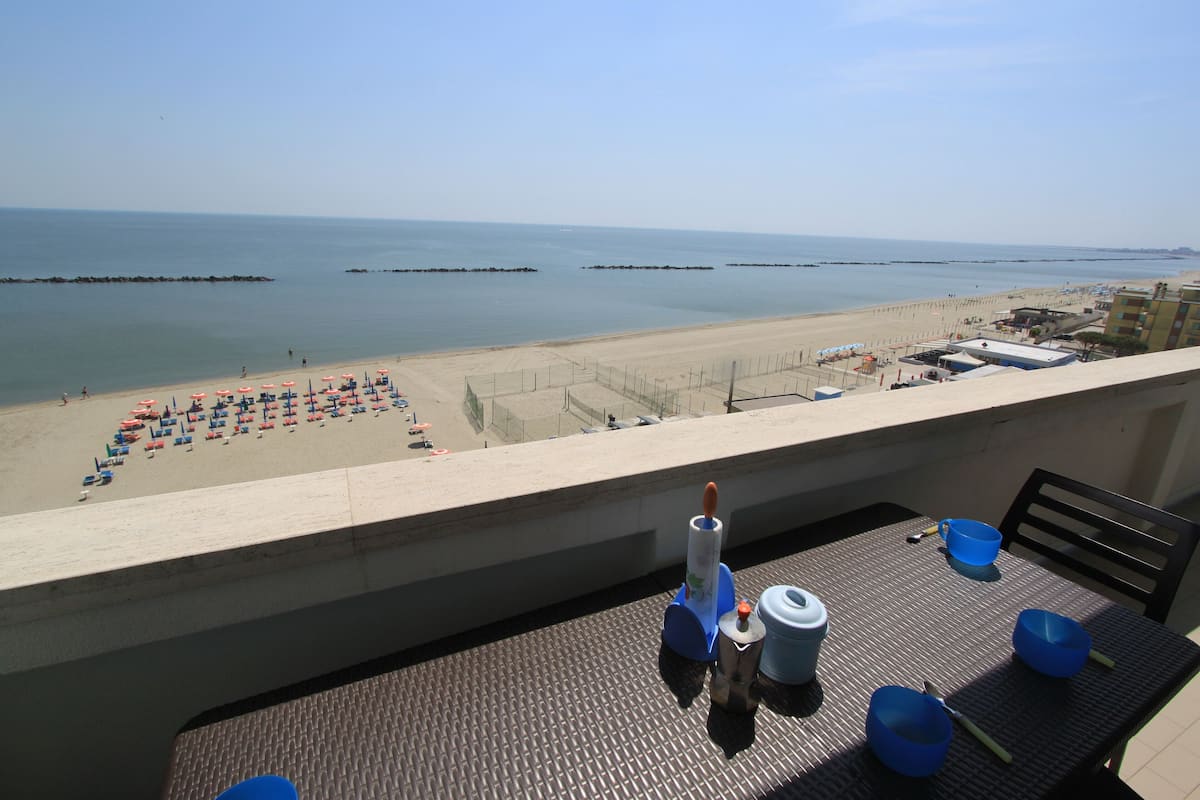 A balcony with a table set for dining, overlooking a beach with umbrellas and a clear view of the sea.