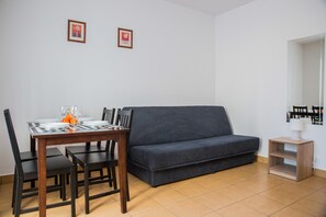 Un salon avec un canapé gris foncé, une table à manger en bois avec des chaises, et une petite table d’appoint en bois sur laquelle se trouve une lampe.