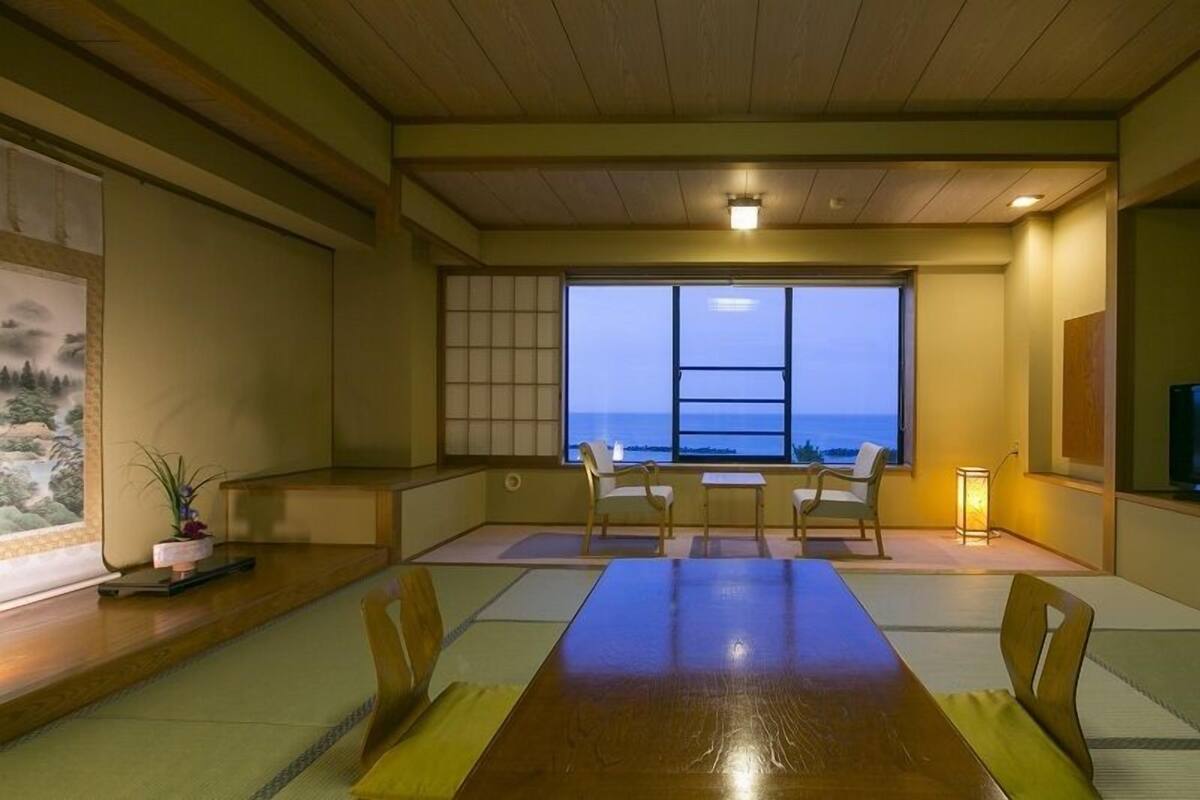 A traditional Japanese-style room with tatami flooring, a low table, chairs, a television, and a large window with a view of the sea.