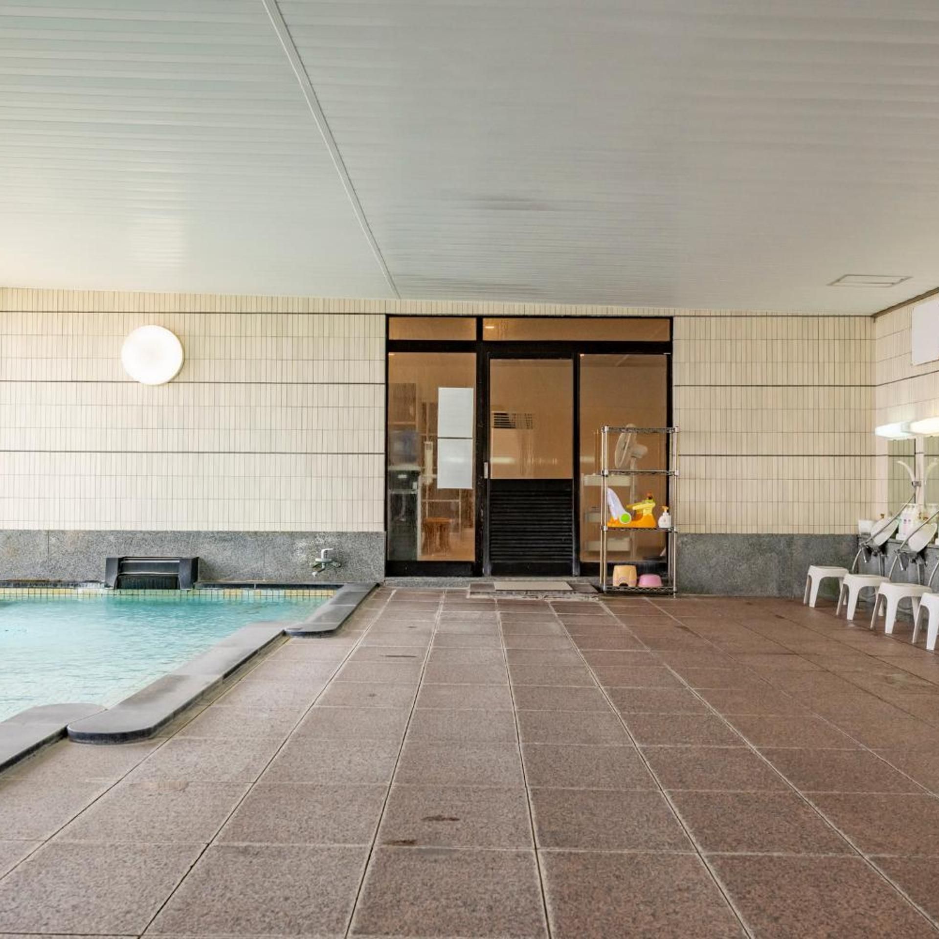 Uma área de piscina com piso de azulejos, porta de vidro que dá para um quarto, e uma fileira de cadeiras brancas à direita.