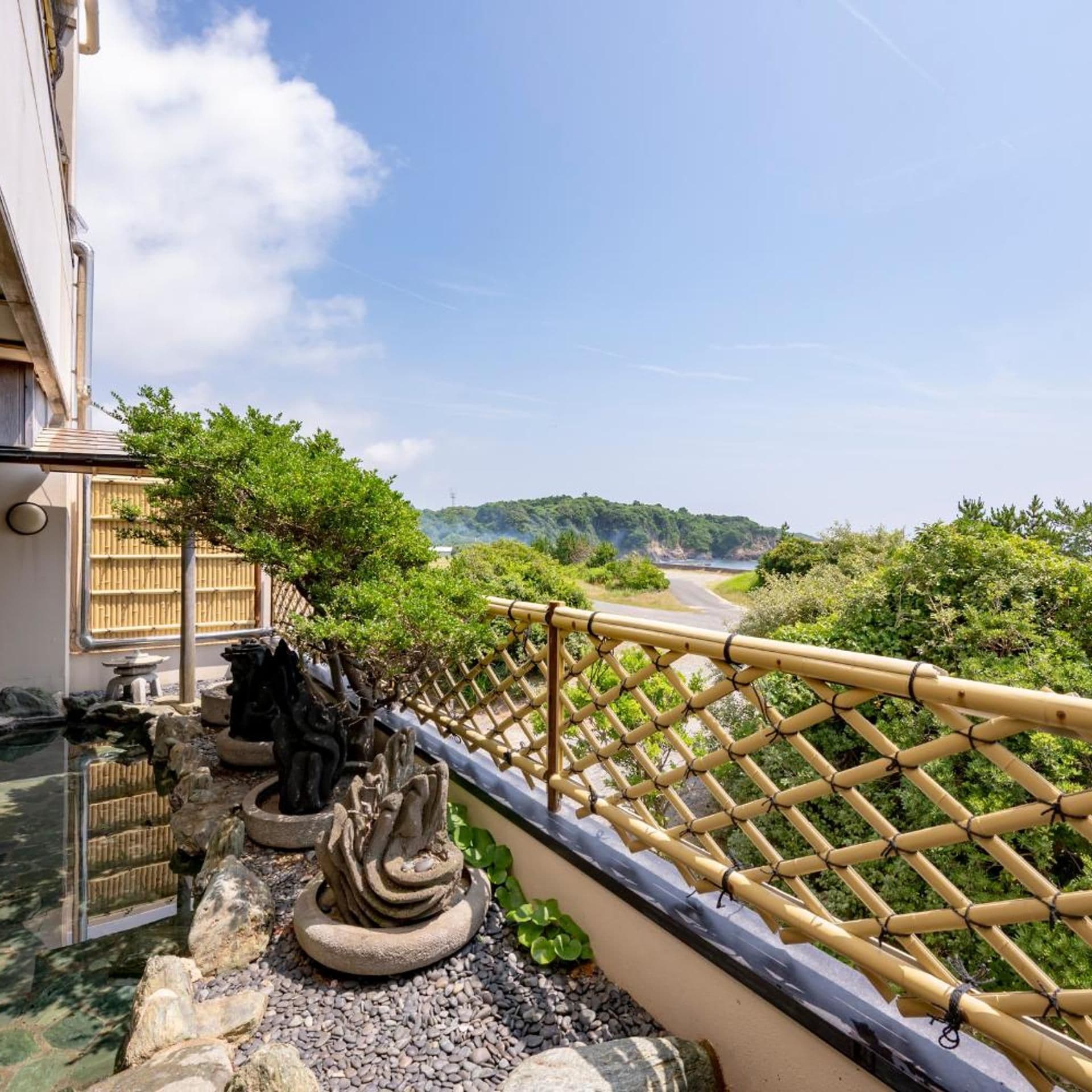 A traditional Japanese garden with a bamboo fence, stone sculptures, and a view of a road and hills.
