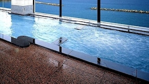 Una piscina interior con vistas al mar, grandes ventanales y suelo de baldosas.