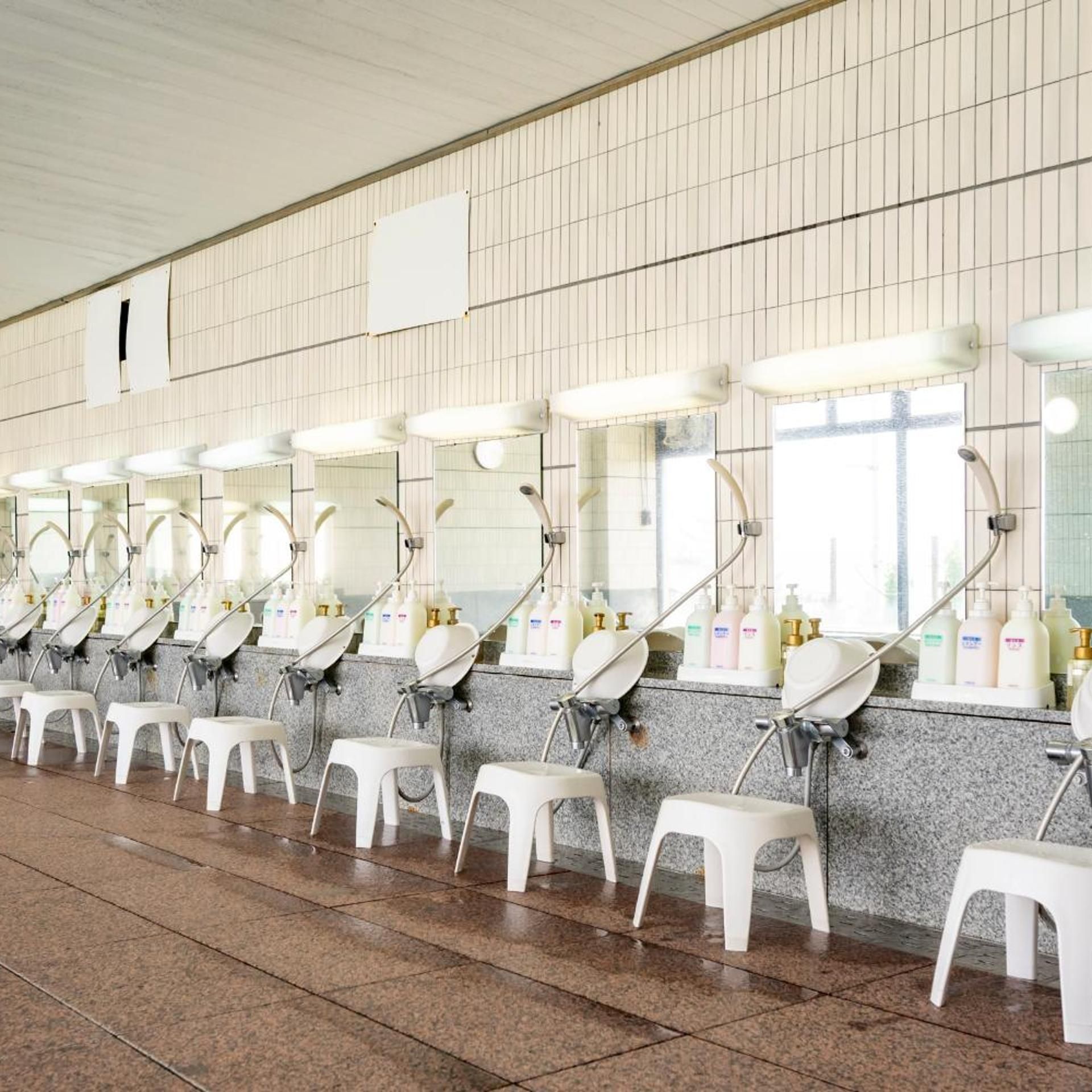 Uma fileira de pias com espelhos e dispensadores de sabão, cada uma com um banquinho na frente.