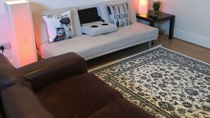 Ein Wohnzimmer mit einer Couch, einem braunen Ledersessel, einem gemusterten Teppich, einem Beistelltisch mit Lampe und einem gerahmten Bild an der Wand.