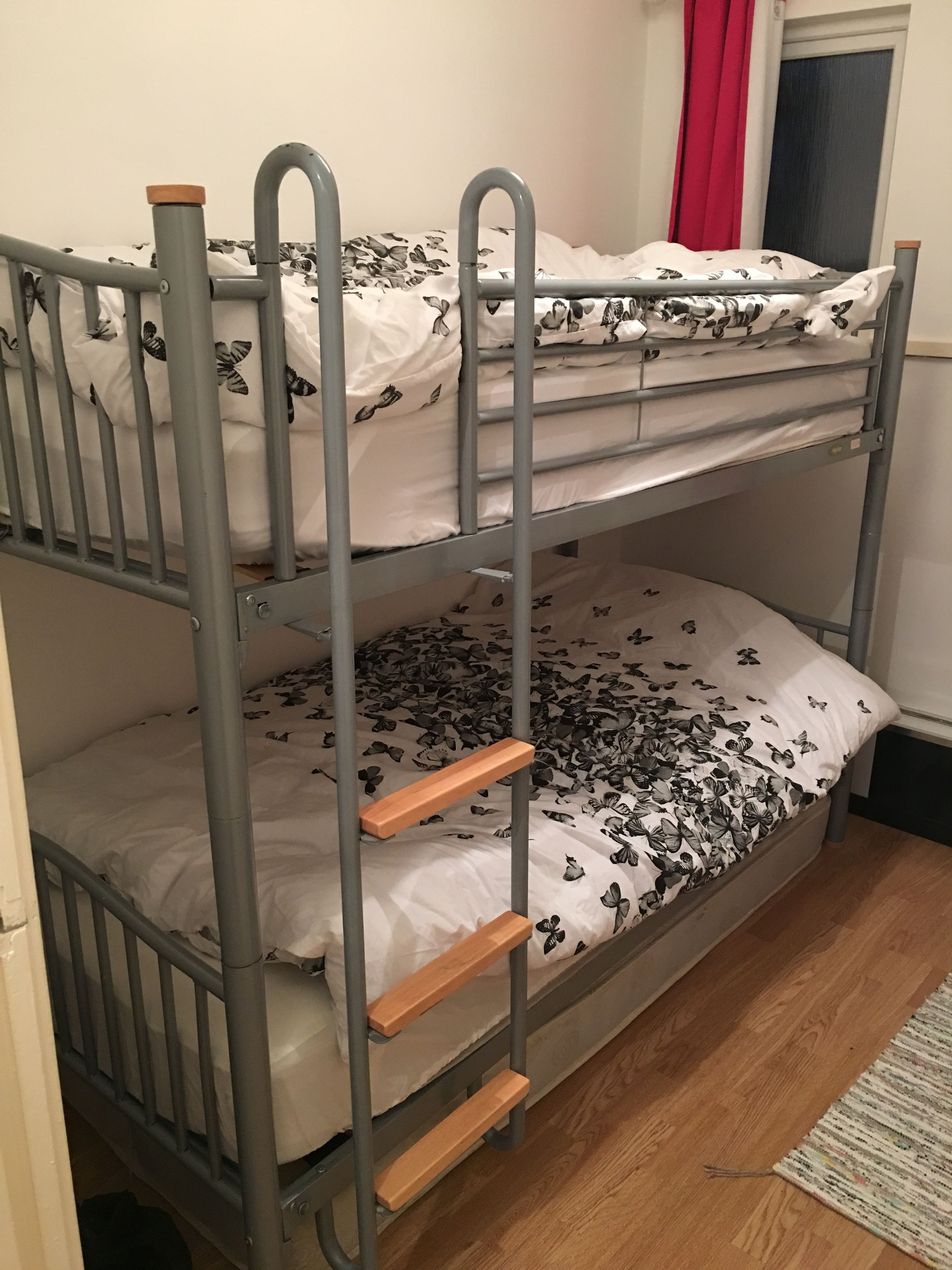 A bunk bed with a floral-patterned bedspread, a wooden ladder, and a window with red curtains.