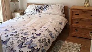 Ein Schlafzimmer mit Bett, Nachttisch, Kommode, Lampe, einem gerahmten Bild an der Wand und einem Fenster mit Vorhängen.