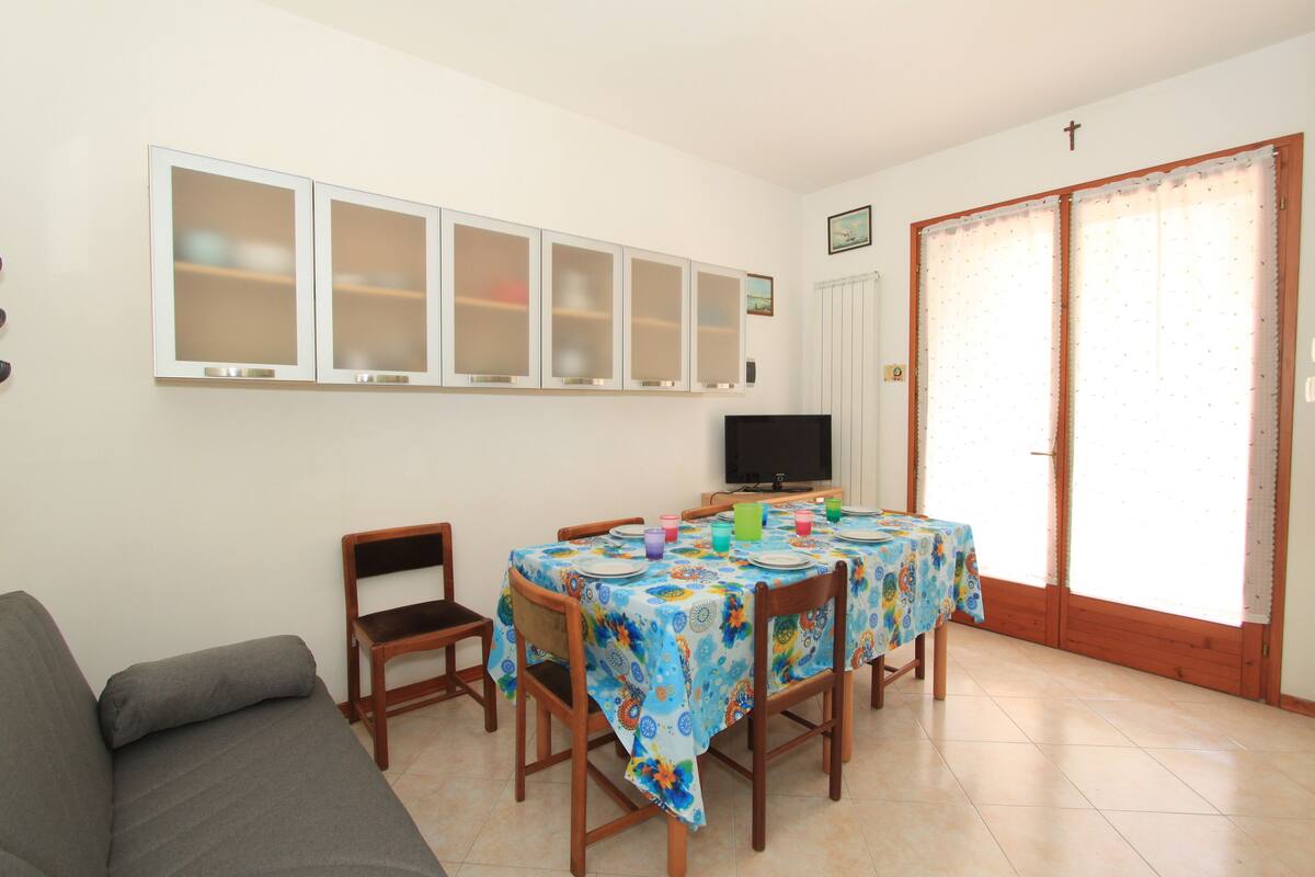 Un comedor con una mesa preparada para cuatro personas, sillas alrededor, un televisor en la pared y una cruz sobre la puerta.