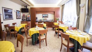 A dining area with round tables covered in yellow tablecloths, wooden chairs, and a kitchen area in the background. - Hotel Brennero (Bassano del Grappa)