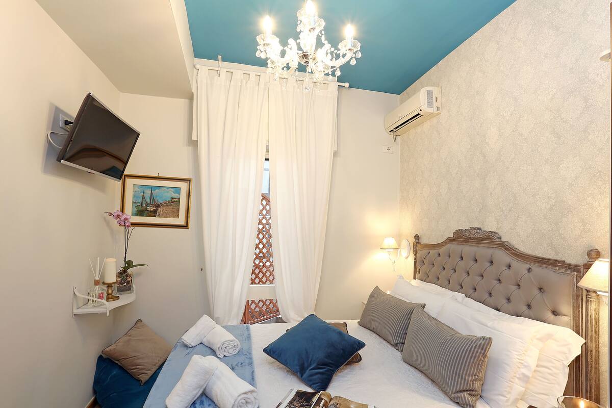 A hotel room with a bed, a mounted TV, a chandelier, and a window with curtains.