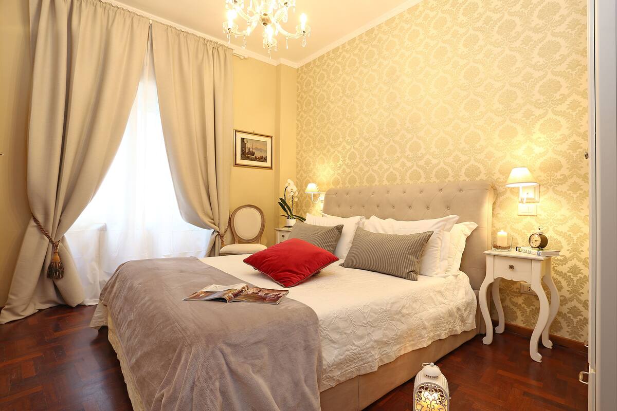 A neatly arranged bedroom with a large bed, a bedside table with a lamp, and a nightstand with a clock.