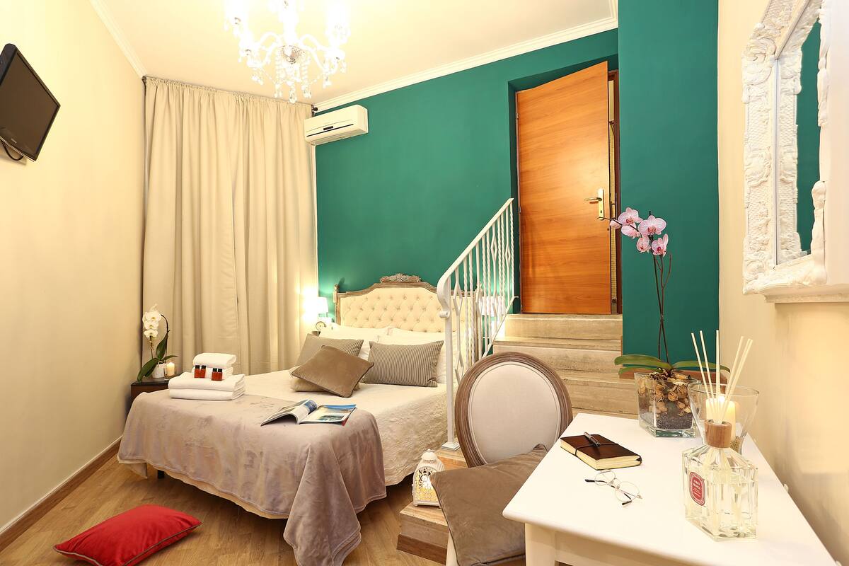 A bedroom with a bed, a bedside table with a mirror, a chair, and a small table with a book and a bottle.