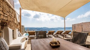 A terrace with a wooden table, lounge chairs, and a view of the sea and mountains. - Mykonos Divino (Mykonos)