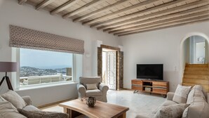 A spacious living room with a large window, a stone fireplace, a flat-screen TV, and a wooden ceiling. - Mykonos Divino (Mykonos)