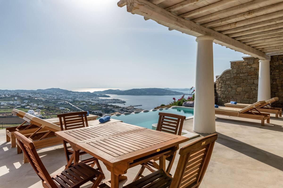 A wooden table and chairs under a pergola with a view of a coastal landscape and a swimming pool.