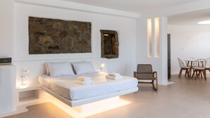 A modern bedroom with a large bed, a stone wall art piece, a wooden bench, and a dining area with chairs. - Mykonos Divino (Mykonos)