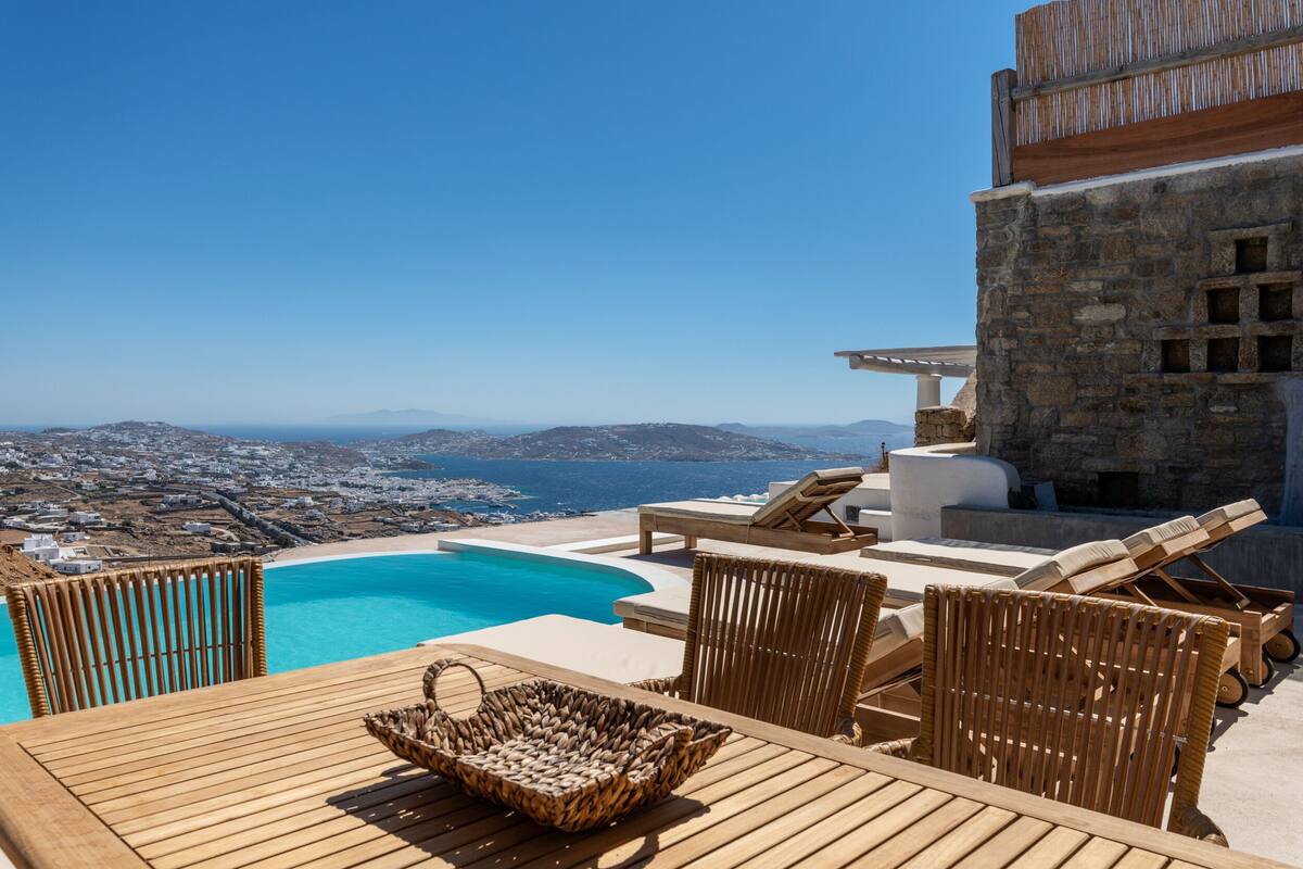 A wooden deck with a view of a beach and ocean, lounge chairs, and a stone wall.