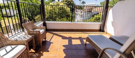 Een overdekt balkon met rieten meubels, een glazen tafel en uitzicht op groen en gebouwen.