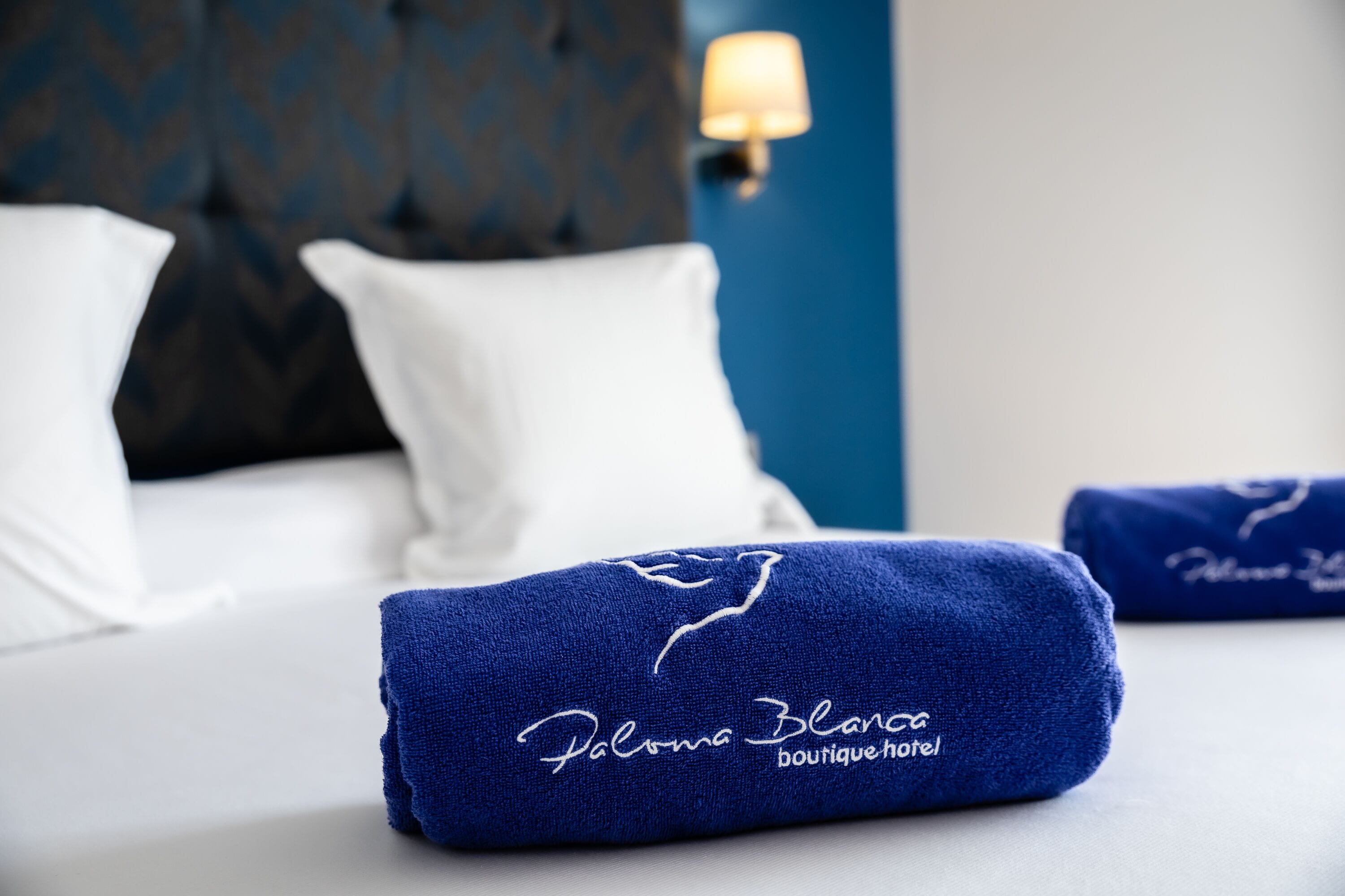A neatly made bed with white linens and a blue towel with embroidery.