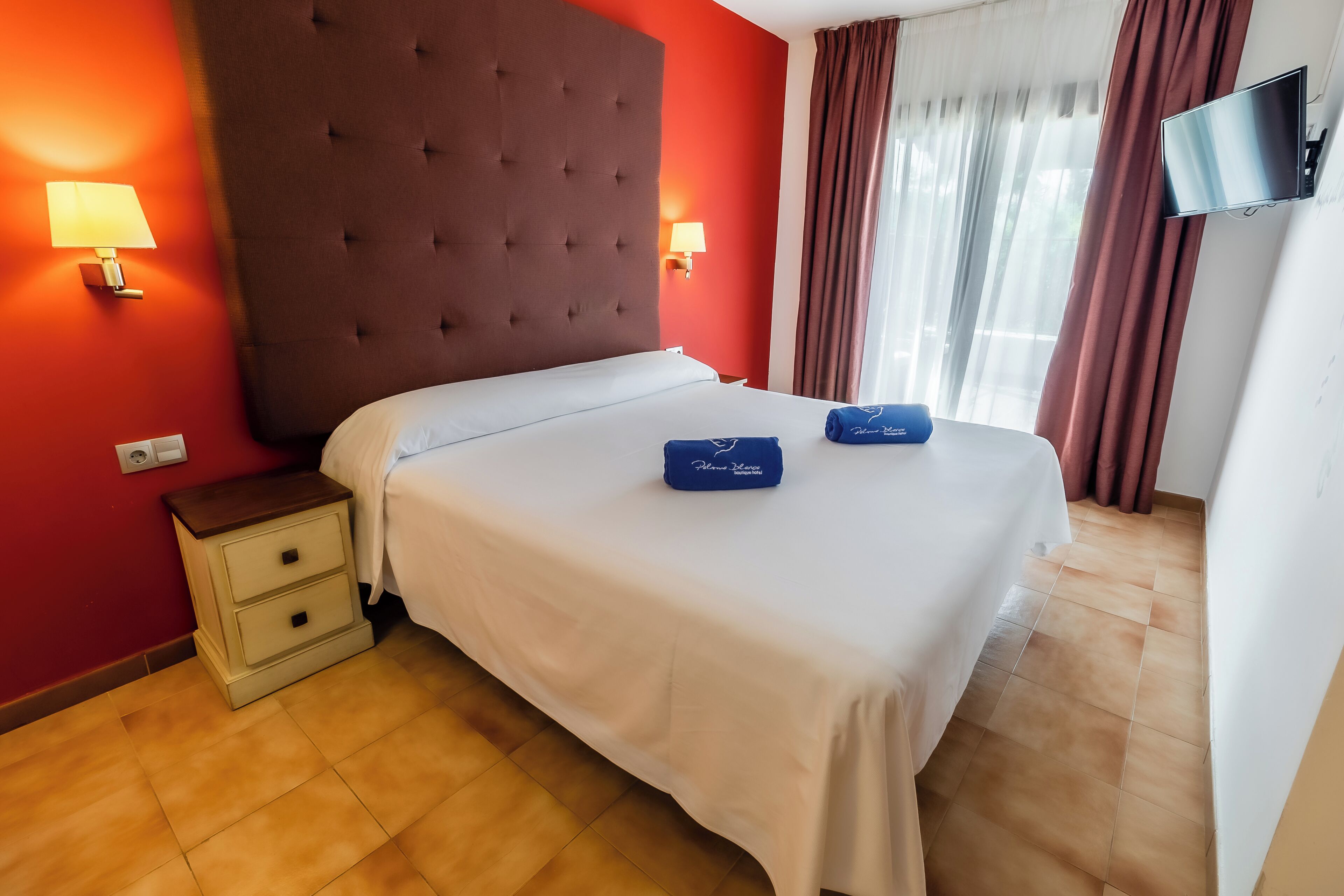 A hotel room with a large bed, two bedside tables, a flat-screen TV, and a window with curtains.