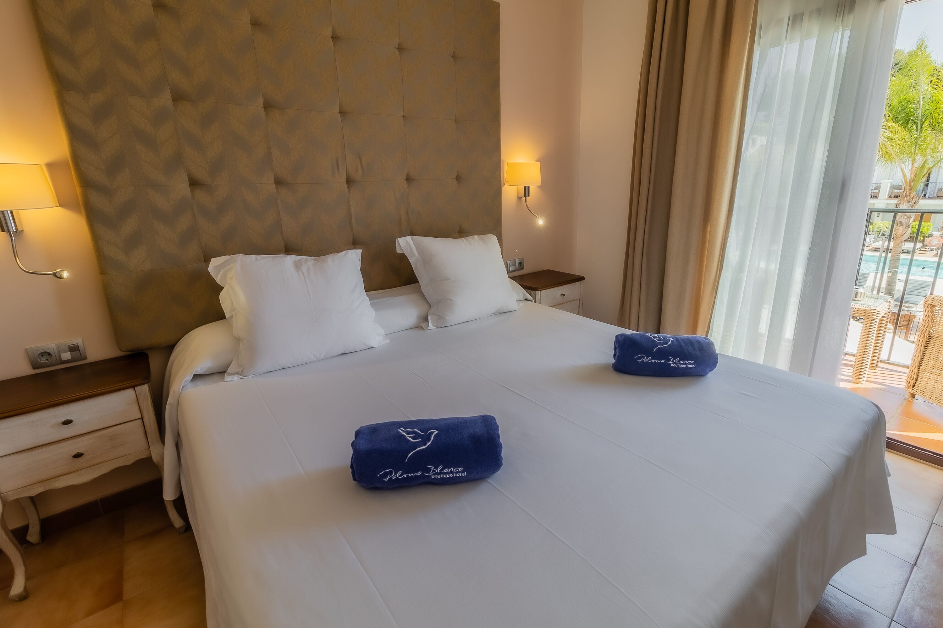 A hotel room with a large bed, two pillows, a nightstand, a lamp, and a view of the outdoors.