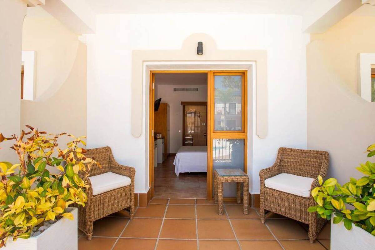 A hallway with two wicker chairs and a small table, leading to a bedroom with a bed and a nightstand.