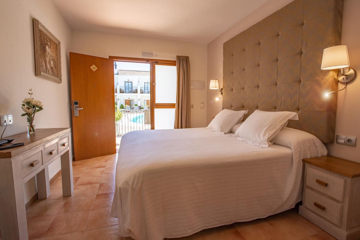 A hotel room with a large bed, a desk with drawers, a bedside table, and a view of a pool through an open door.