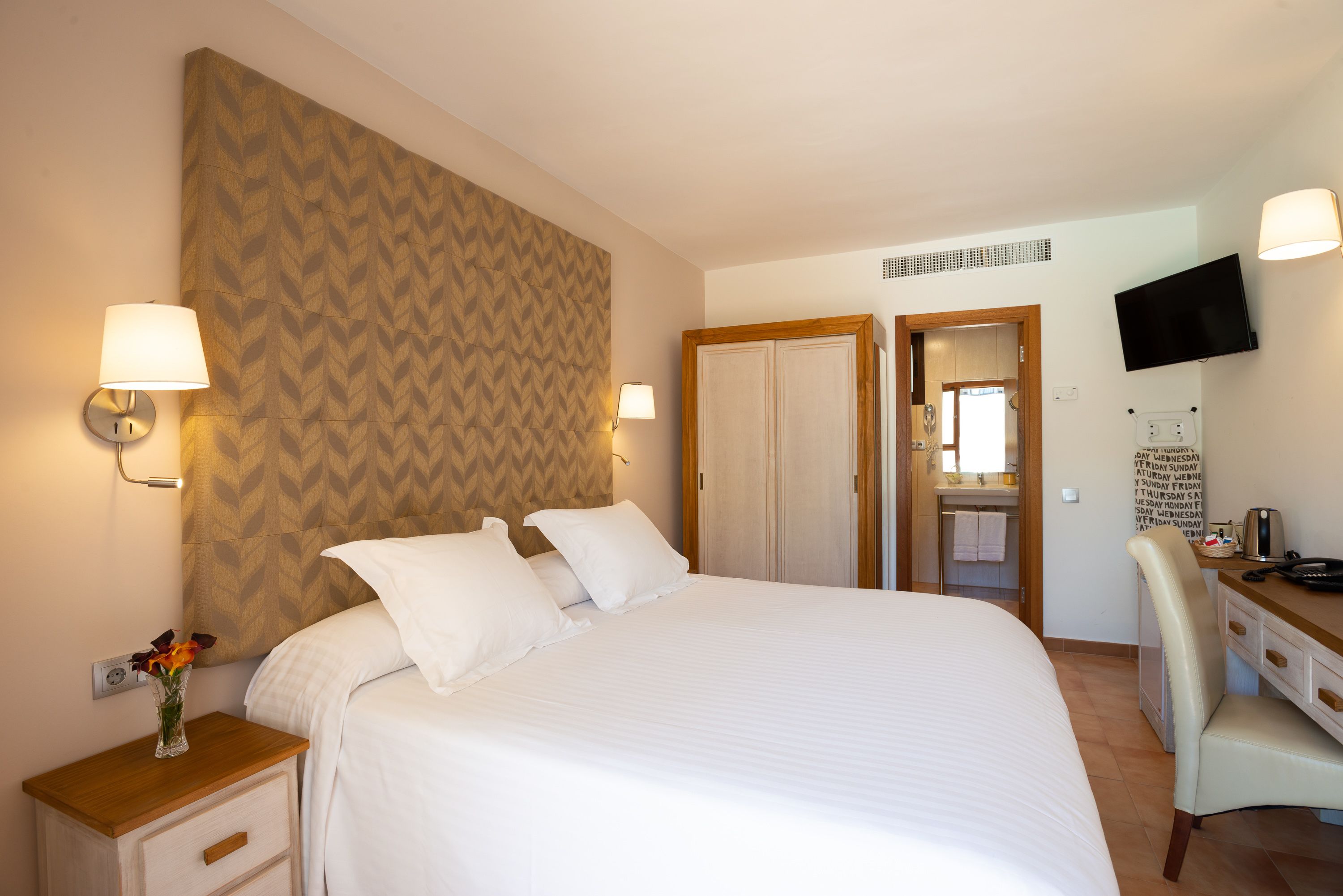 Habitación de hotel con una cama grande, dos lámparas de noche, una mesita de noche de madera con un florero, un escritorio con una silla, un televisor y un armario.