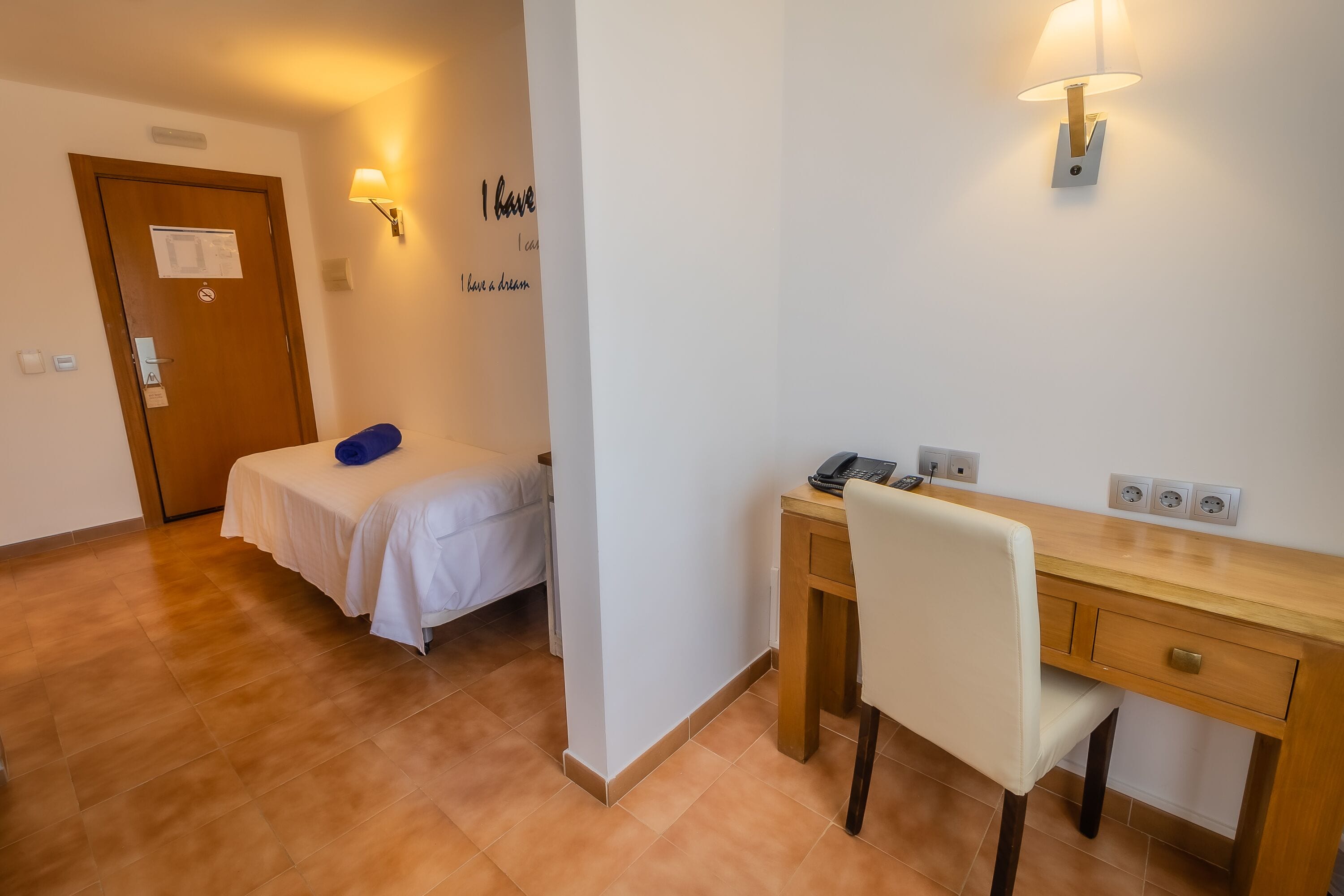 A hotel room with a bed, a desk, a chair, a lamp, and a wall-mounted light.