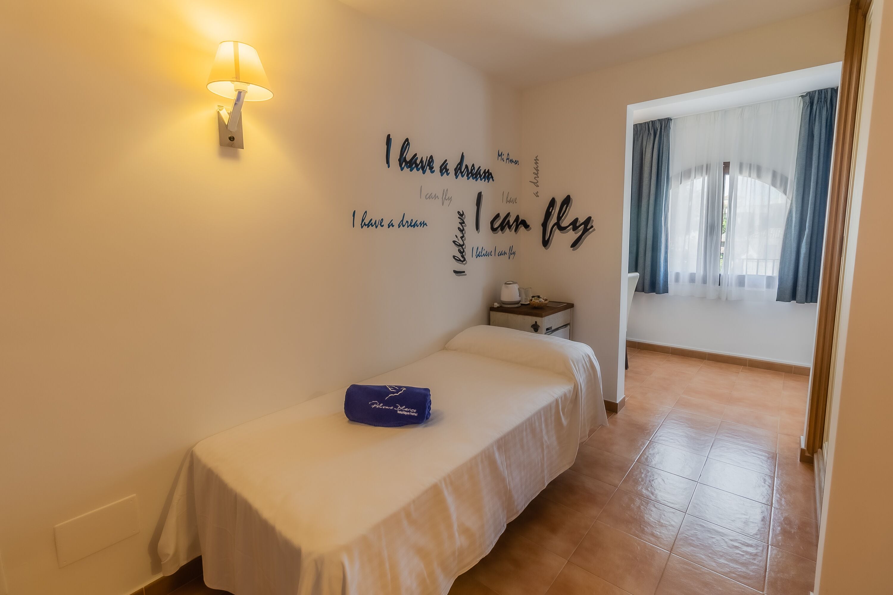 A bedroom with a bed, a wall-mounted lamp, a nightstand, and a window with curtains.