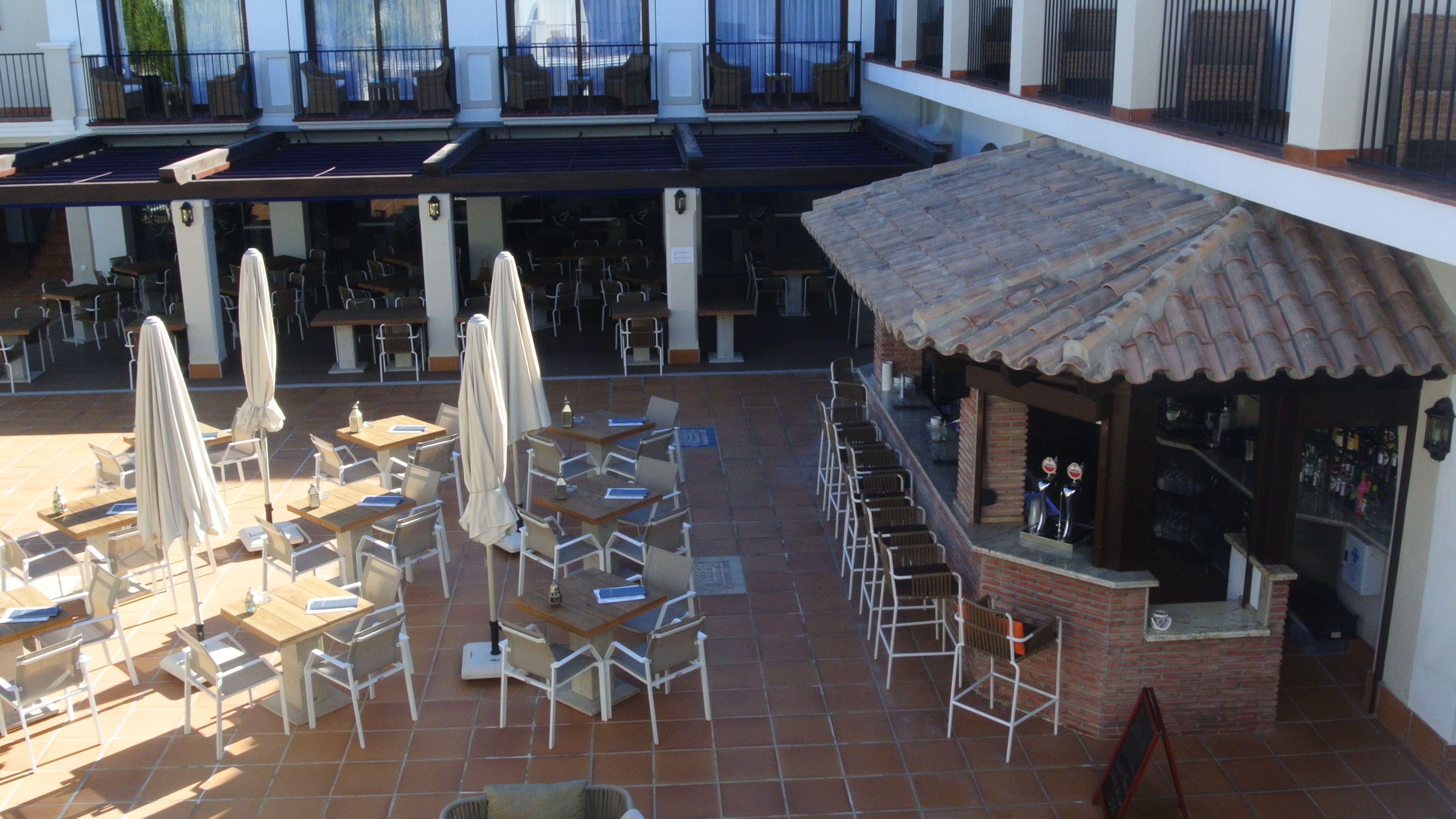 An outdoor patio area with tables and chairs, umbrellas, and a bar area with a tiled roof.
