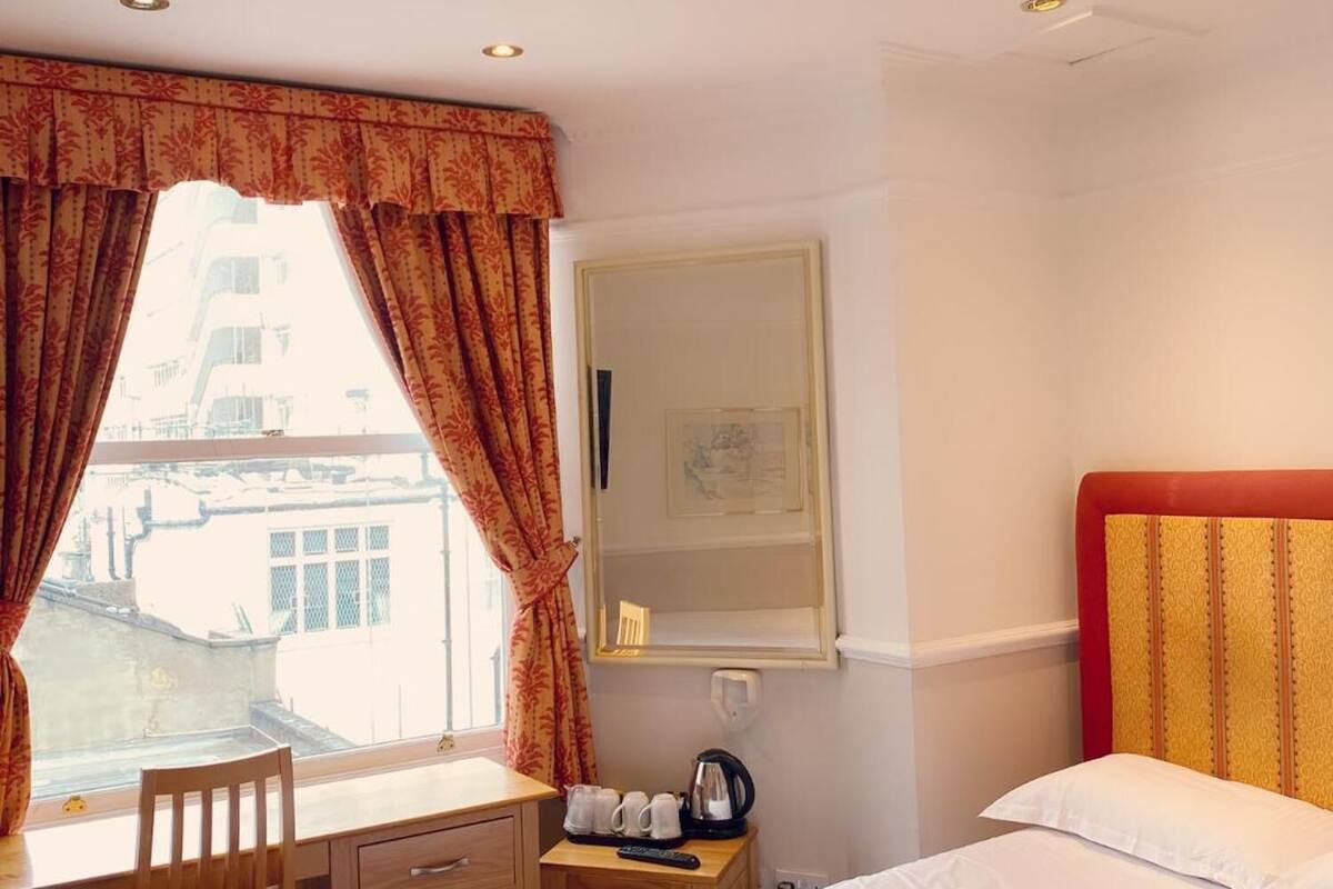 A hotel room with a bed, a desk with a chair, a small table with a kettle, and a window with curtains.