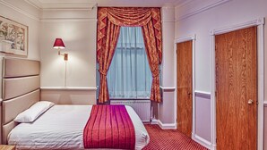A hotel room with a bed, a nightstand, a lamp, a window with curtains, a radiator, and a wooden door. - Hyde Park Radnor Hotel (London)