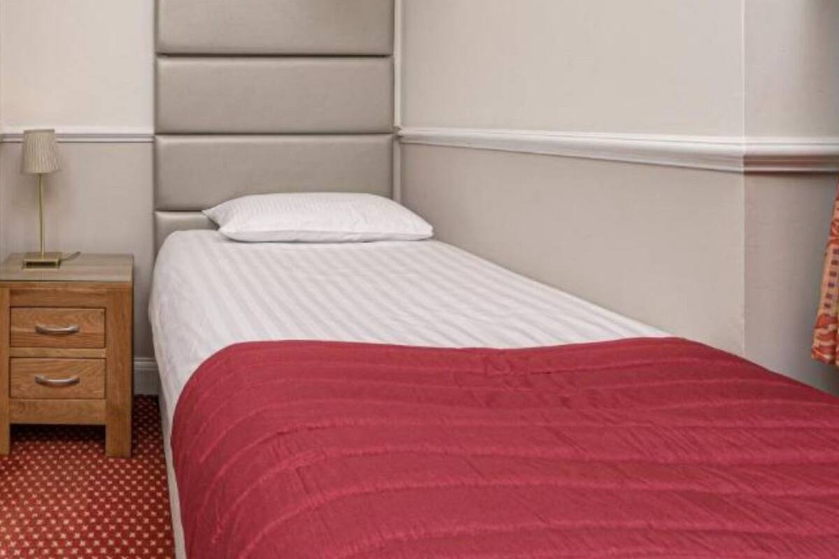 A single bed with a white mattress and a red blanket, a wooden nightstand with drawers, and a lamp.