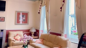 A room with two beige sofas, a wooden coffee table, a chandelier, and a large window with curtains. - Hyde Park Radnor Hotel (London)