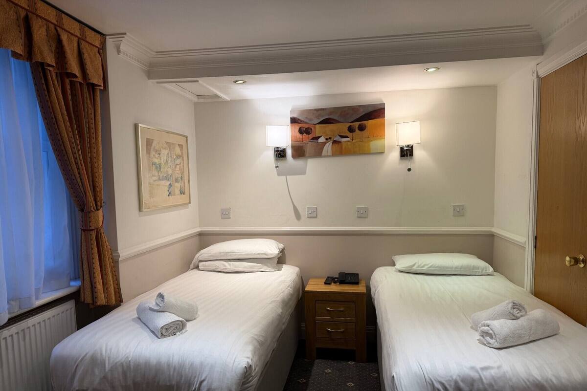A hotel room with two single beds, a nightstand, a painting, and a window with curtains.