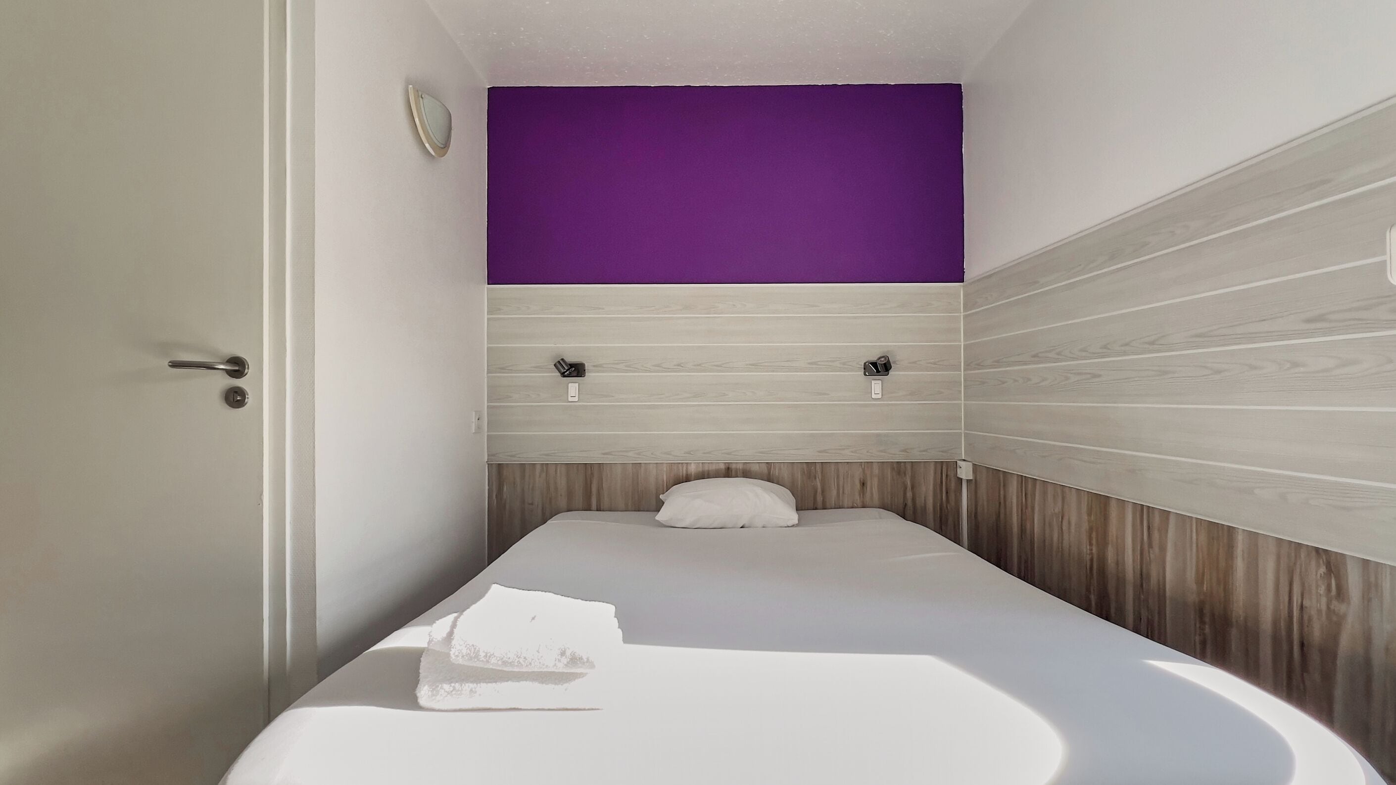 Une petite chambre avec un lit simple, un mur lambrissé en bois, un mur d’accent violet et un lit blanc.