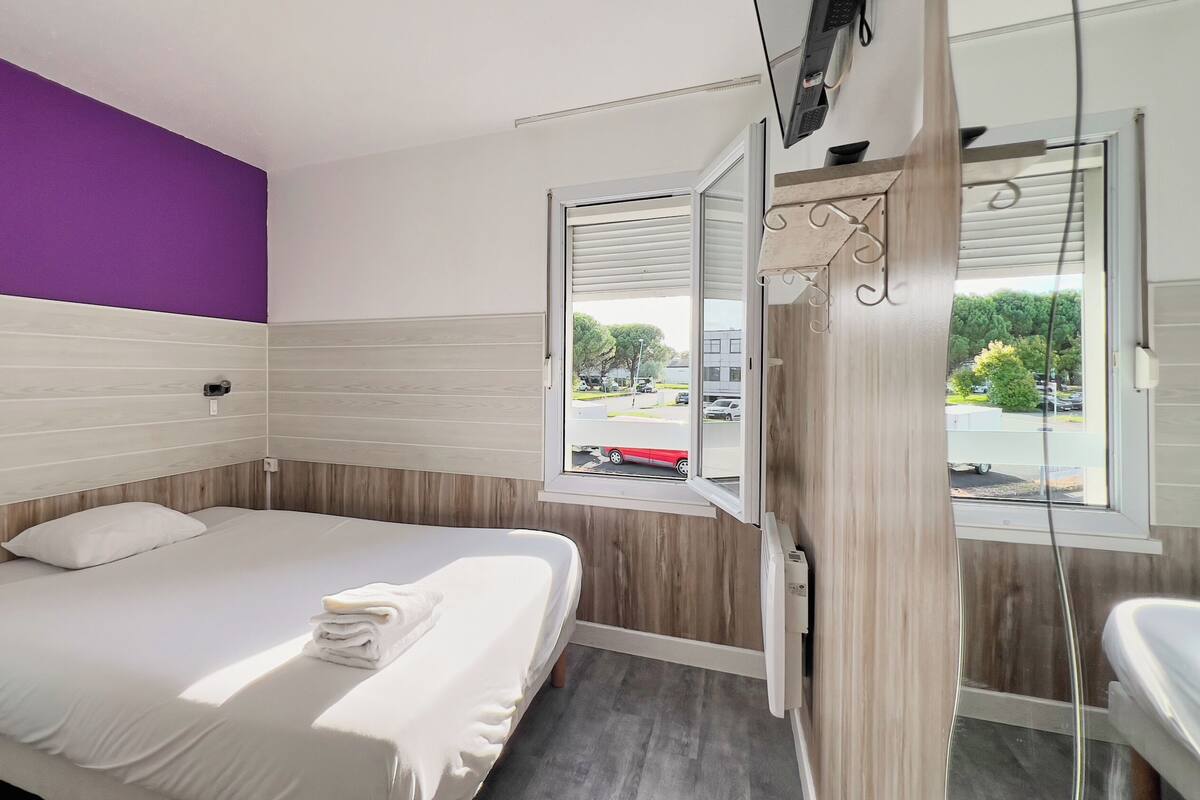 Cama bem arrumada com lençóis brancos em quarto com parede de destaque roxa e janela com persianas.