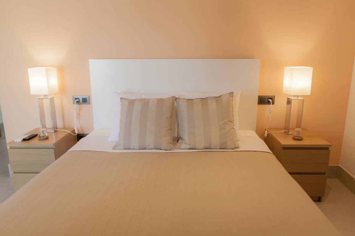 A neatly made bed with two pillows, two bedside tables with lamps, and a nightstand with a remote control.
