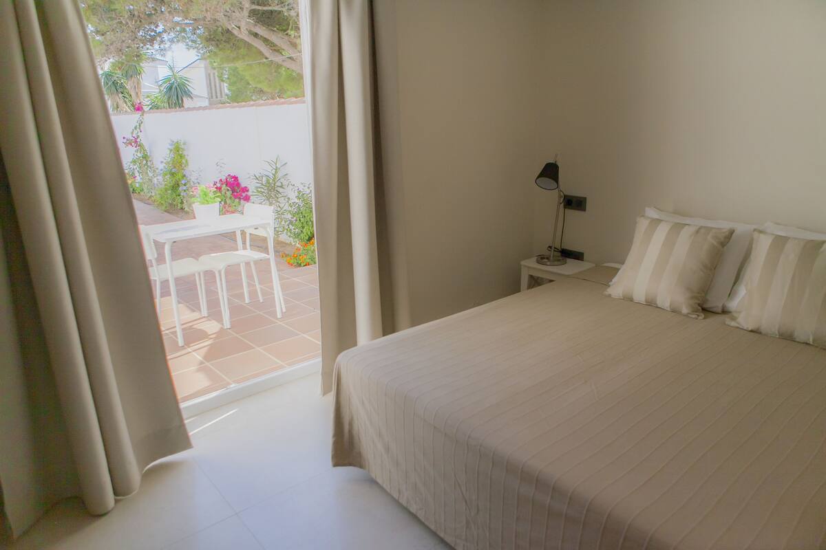 A bedroom with a large bed, a bedside table with a lamp, and a view of an outdoor patio with a table and chairs.