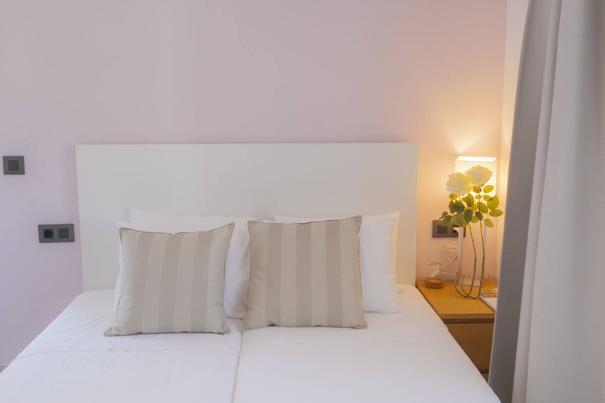 A neatly made bed with two pillows, a bedside table with a lamp, and a wall-mounted thermostat.